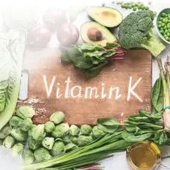 Grünes Gemüse wie Spinat, Brokkoli und Rosenkohl, das auf einem Holzbrett mit der Aufschrift "Vitamin K" arrangiert ist, zeigt, was Vitamin K ist.